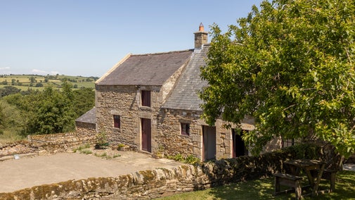 An 18th century stone cottage with a farmyard in front and surrounded by trees and green fields